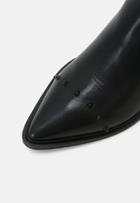 Scarpa in pelle nera a punta con piccoli chiodi tondi e a forma di cono neri sulla parte superiore anteriore, su sfondo bianco.