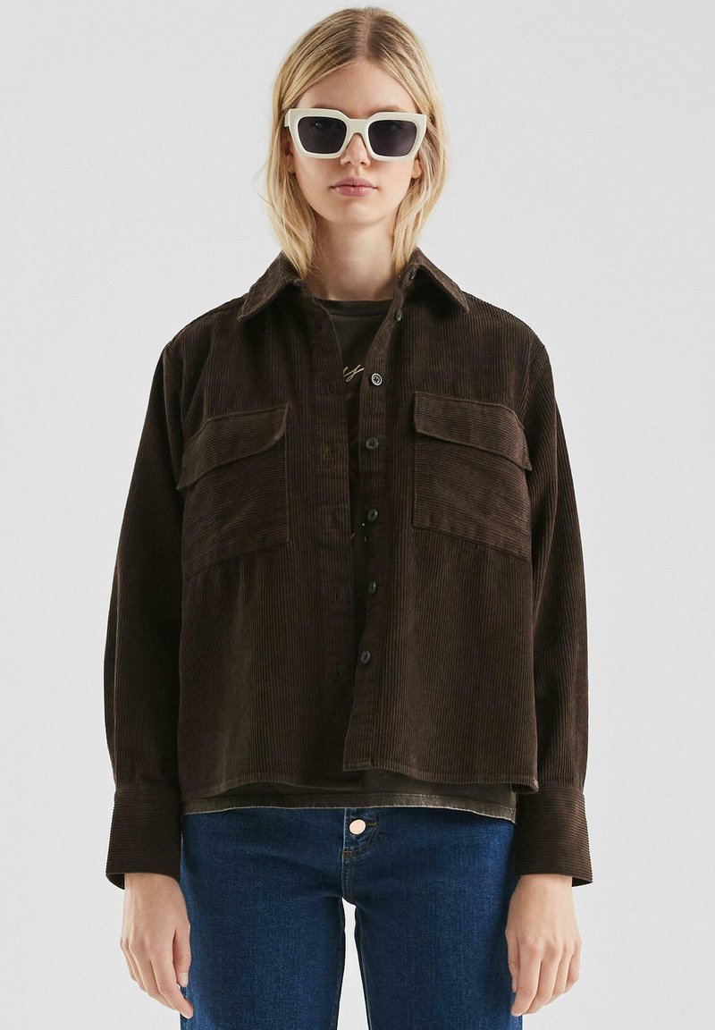 Braune Cordjacke mit zwei Brusttaschen, Knopfverschluss und Kragen. Wird über einem dunklen Oberteil getragen und mit blauen Jeans kombiniert.