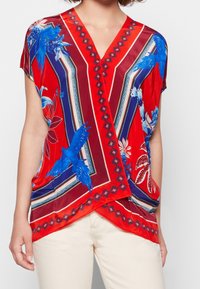 Blouse rouge à motifs avec des dessins d'oiseaux bleus, un col en V et des manches courtes. Le tissu a une finition lisse et brillante et offre une coupe décontractée.