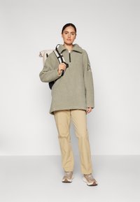 Pullover in pile verde oliva con colletto a mezza zip, forma rilassata e dettaglio a toppa sulla manica, abbinato a pantaloni beige e sneakers chiare.