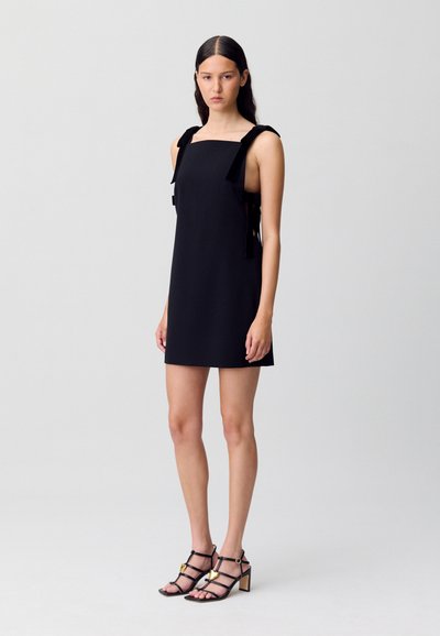 Donna in piedi che indossa un mini abito nero senza maniche con spalline annodate e sandali con tacco alto neri e cinturini con accenti dorati.