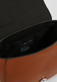 Ruskea keinonahkainen laukku, jossa on sileä pinta, musta kangasvuori ja näkyvä logotunnus. Suunnittelussa on magneettisuljin.
