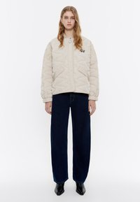 Giacca trapuntata beige con colletto tondo, dotata di tasche laterali e un dettaglio ricamato, abbinata a jeans larghi scuri e scarpe nere.