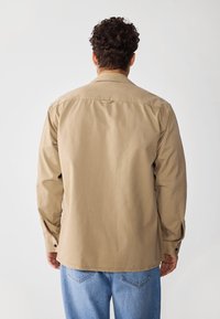 Camisa de algodón beige de manga larga, con yugo trasero y puños abotonados, combinada con unos vaqueros de color azul claro, vista desde la parte trasera.