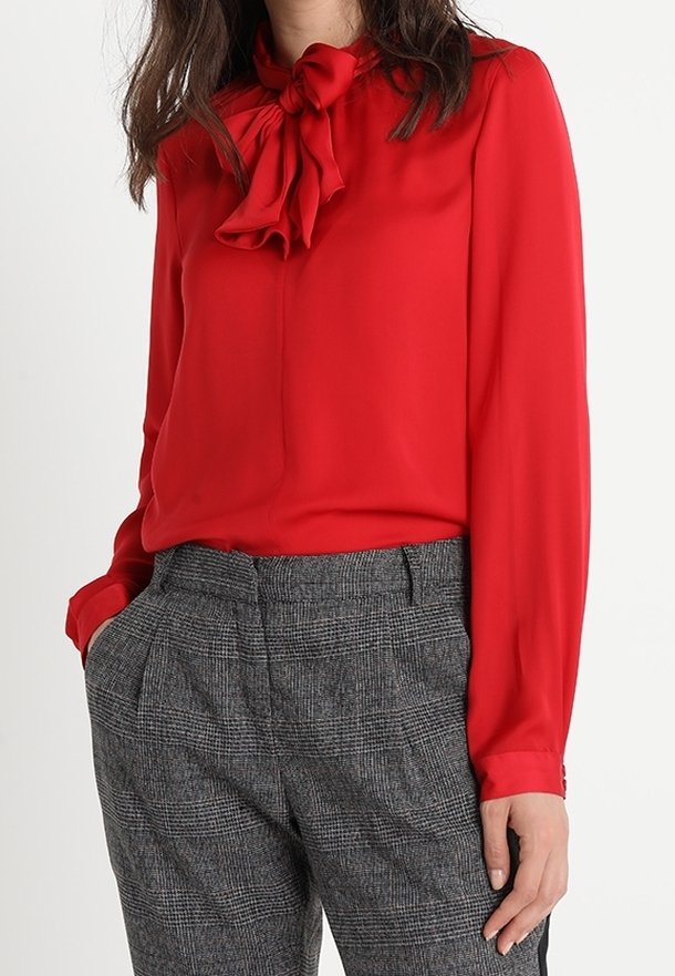 Blouse rouge à manches longues avec un détail de nœud au col, associée à un pantalon tailleur gris à carreaux avec une finition texturée et des poches latérales.