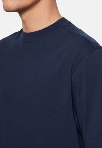 Tricou din bumbac de culoare albastru marin, cu guler rotund clasic și mâneci scurte. Prezintă o cusătură plată și o textură netedă. Fără modele sau accente.