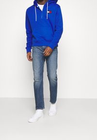 Felpa blu royal con tasca frontale e colletto con zip, abbinata a jeans blu chiaro e sneakers bianche. Presenta un logo sul petto.