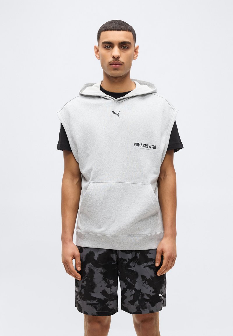 Joven con sudadera sin mangas gris de Puma sobre una camiseta negra y pantalones cortos de camuflaje negro y gris de Puma, de pie contra un fondo liso.