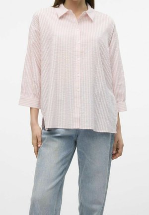 Kobieta ubrana w luźną, jasnoróżową koszulę w białe paski z guzikami, rękawy do łokcia, oraz jasnoniebieskie dżinsy.