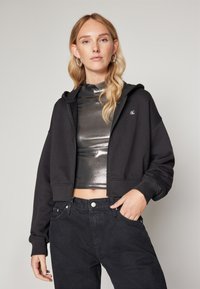 Haut à capuche noir court avec une fermeture éclair à l'avant, des manches longues et un logo texturé. Porté par-dessus un haut argent métallique et associé à un jean noir taille haute.