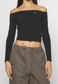 Schwarzes, schulterfreies Rippenoberteil mit langen Ärmeln, das über ein dezentes Logo verfügt. Kombiniert mit lockeren, khakifarbenen Hosen.