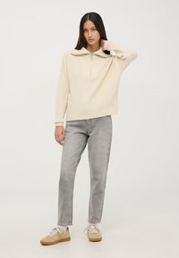 Sweter w kolorze kremowym, prążkowany, z ćwierć zamkiem i luźnym krojem, w połączeniu z jasnoszarymi dżinsami o prostym kroju oraz beżowymi trampkami.