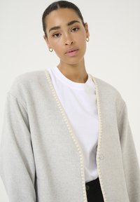 Cardigan grigio realizzato in materiale morbido con rifinitura crema, chiusura a bottoni e vestibilità rilassata, indossato sopra una t-shirt bianca.