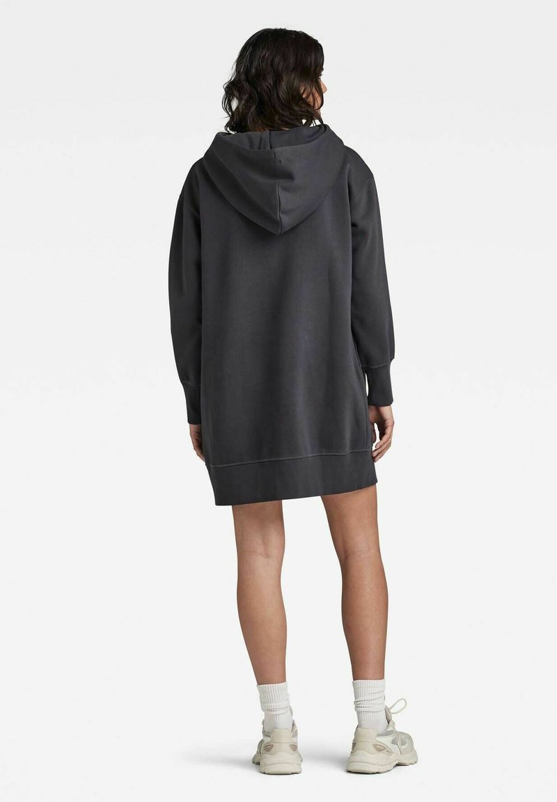 Femme aux cheveux bouclés foncés portant une longue robe sweat à capuche gris foncé, des chaussettes blanches et des baskets de couleur claire, vue de dos.