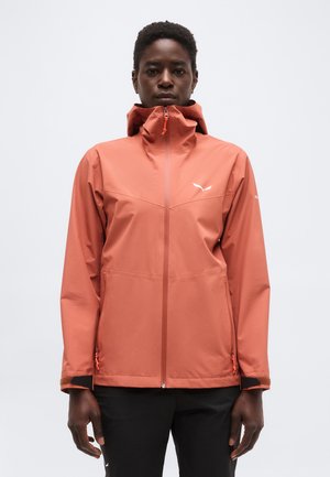 Άτομο που φοράει αδιάβροχο μπουφάν με κουκούλα σε ροζ-σομόν χρώμα, με φερμουάρ μπροστά, και μαύρο παντελόνι, με φόντο απλό λευκό.