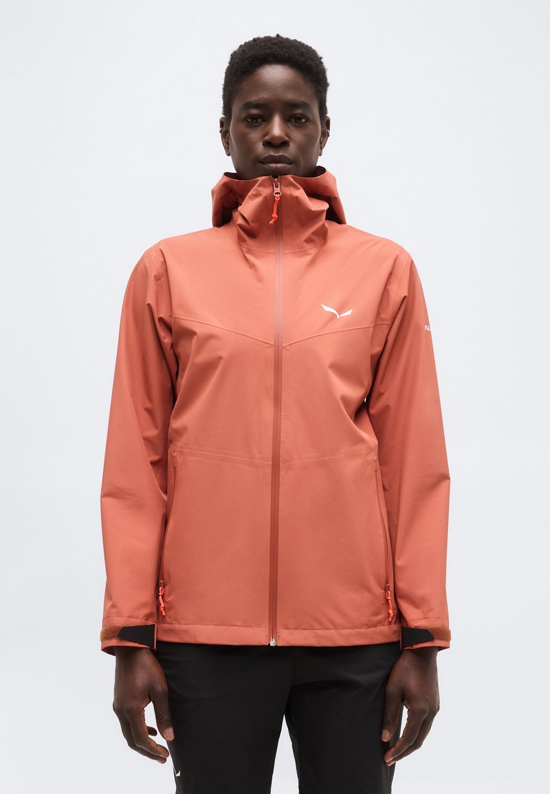 Άτομο που φοράει αδιάβροχο μπουφάν με κουκούλα σε ροζ-σομόν χρώμα, με φερμουάρ μπροστά, και μαύρο παντελόνι, με φόντο απλό λευκό.