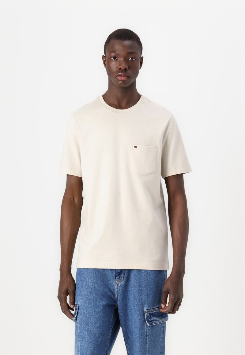 Beige cotton t-shirt with a crew neckline and short sleeves, featuring a small chest pocket and the brand's logo in color. Worn with blue jeans.