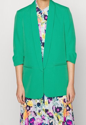 Persoon draagt een felgroene blazer met driekwart mouwen over een meerkleurige bloemrijke jurk met knopen.