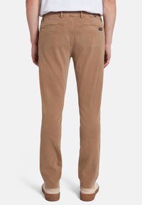 Pantaloni in cotone marrone con taglio dritto, due tasche posteriori e una texture sottile. Indossati con scarpe di colore chiaro, in piedi contro uno sfondo bianco.