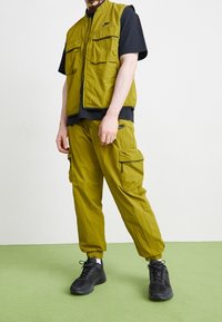 Olive green vest and cargo pants made of lightweight fabric, featuring multiple pockets and elastic cuffs, paired with black sneakers.