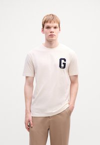Jeune homme aux cheveux courts portant un t-shirt écru avec un grand écusson "G" bleu marine et un pantalon beige, se tenant devant un fond blanc uni.