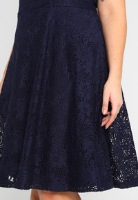 Navy lace dress featuring a floral pattern with layered texture. The skirt is A-line, showcasing intricate cut-out designs at the hem.