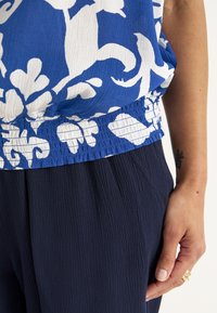 Blå og hvid blomstret top med en samlet elastisk talje, parret med mørkeblå strukturerede bukser. Nærbillede af beklædningsdetaljer.