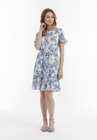 Floral Kleid in Hellblau und Creme mit kurzen Rüschensäumen, geraffter Taille und ausgestelltem Saum, kombiniert mit grauen Riemchensandalen.