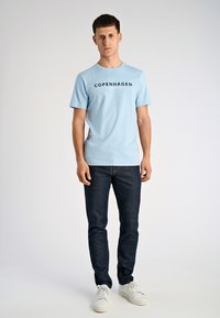 Maglietta in cotone azzurro chiaro con le scritte "COPENHAGEN" e "LINDBERGH" in nero, abbinata a jeans di denim scuro e sneakers bianche. Design semplice.