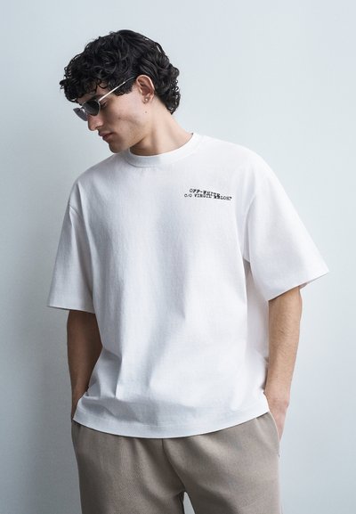 Homme aux cheveux bouclés et foncés portant des lunettes de soleil noires, un T-shirt blanc oversized avec un petit texte, et un pantalon beige, debout les mains dans les poches.