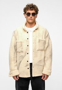 Beige fleece jack met een knoopsluiting aan de voorkant, twee borstzakken en een kraag. Draagt over een wit shirt en zwarte broek.