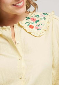 Chemise jaune à boutons avec un col froncé et une broderie florale colorée. Texture lisse avec une coupe décontractée et un éclat subtil.