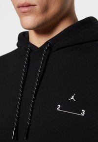 Homme portant un sweat à capuche noir avec des cordons noirs et blancs et un petit logo Jumpman blanc avec "2 3" imprimé sur la poitrine.