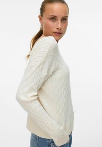 Kremowy dzianinowy sweter w teksturowany wzór diamentowy, z dopasowanymi mankietami i okrągłym dekoltem. Noszony na niebieskich dżinsach.