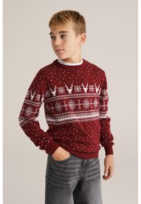 Piros kötött pulóver fehér hópehely és rénszarvas mintával, kerek nyakkivágással és bordázott mandzsettával. Szürke farmerrel viselve.