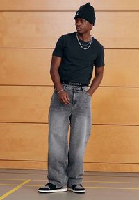 Μαύρο πουκάμισο με κοντά μανίκια, γκρι φαρδιά τζιν, μαύρο σκουφί και μαύρα αθλητικά παπούτσια με λευκές σόλες. Απλός σχεδιασμός, άνετη εφαρμογή και ελάχιστες λεπτομέρειες.