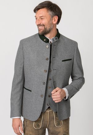 Mann trägt traditionelle graue Wolljacke mit dunkelgrünem Besatz und bestickte braune Lederhose, lächelnd und nach links schauend.