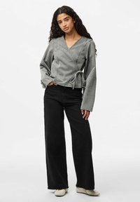 Chaqueta gris con manga larga y detalle de lazo, combinada con pantalones de pierna ancha negros y zapatos de color claro. Textura de tela suave.
