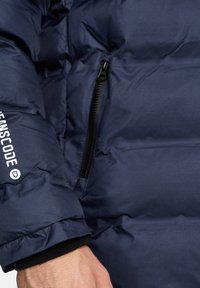Chaqueta acolchada azul marino con superficie texturizada, que presenta un bolsillo con cremallera negra y una marca blanca en la manga, con puños elásticos.