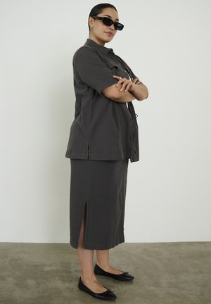 Woman wearing dark gray short-sleeve jacket and matching skirt with side slit, black flats, and black sunglasses, standing with arms crossed.