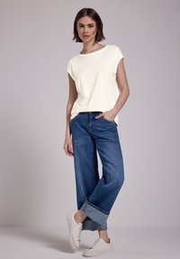 Cremefarvet kortærmet t-shirt kombineret med vide blå denim jeans. Modellen bærer hvide sneakers. Enkel og afslappet design, minimal detaljering.