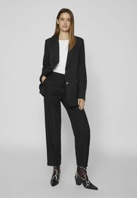 Blazer nero su misura con bottoni, camicia bianca sotto, pantaloni neri e stivaletti neri alla caviglia con dettagli bianchi. Design minimalista.