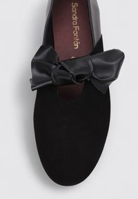 Zapato de ante negro con punta redonda, que cuenta con un gran lazo de satén negro en la parte frontal. La plantilla tiene texto de marca en dorado.