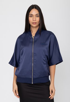 Vrouw met lang donker haar die een marineblauwe satijnen bomberjack met korte mouwen en een gouden ritssluiting draagt, gecombineerd met een zwarte rok.