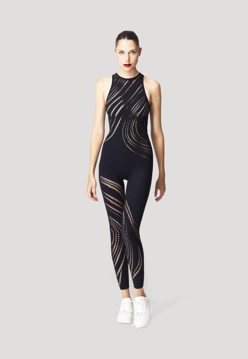 Tuta nera senza maniche con motivi ritagliati, tono della pelle a contrasto, texture liscia. Abbinata a sneakers bianche.