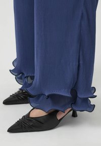 Pantalons navy plissés et fluides avec un ourlet ondulé, associés à des escarpins noirs pointus avec des détails en cuir froncé et un petit talon.