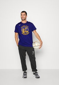 Camiseta azul marino con el logo de "Golden State Warriors" y acentos amarillos, combinada con pantalones de chándal negros, sosteniendo un balón de baloncesto, y usando zapatillas.
