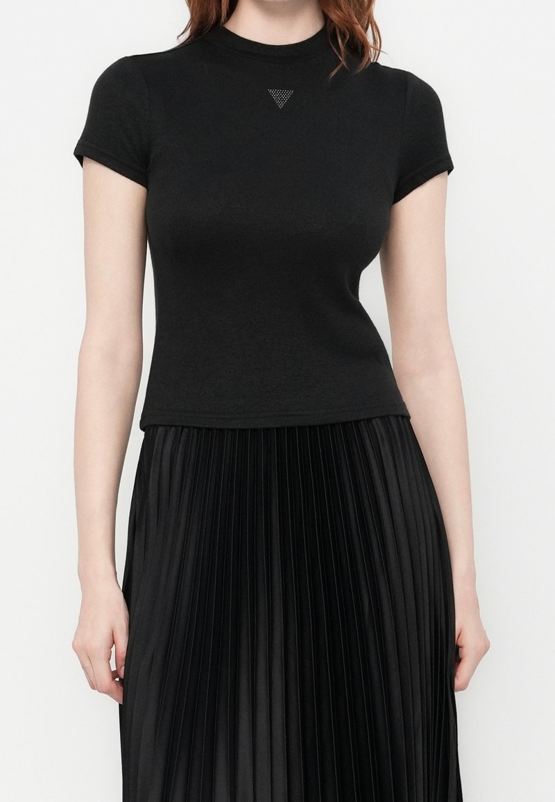 Femme portant un haut noir ajusté à manches courtes avec un petit détail triangulaire et une jupe plissée noire taille haute, debout devant un fond uni.