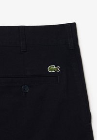 Pantaloni blu scuro in cotone con texture liscia. Presentano un logo di coccodrillo ricamato verde e un bottone sulla vita.