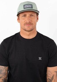 Casquette vert clair avec un logo en patch noir, portée avec un t-shirt noir à manches courtes comportant un petit accent logo blanc sur la poitrine.